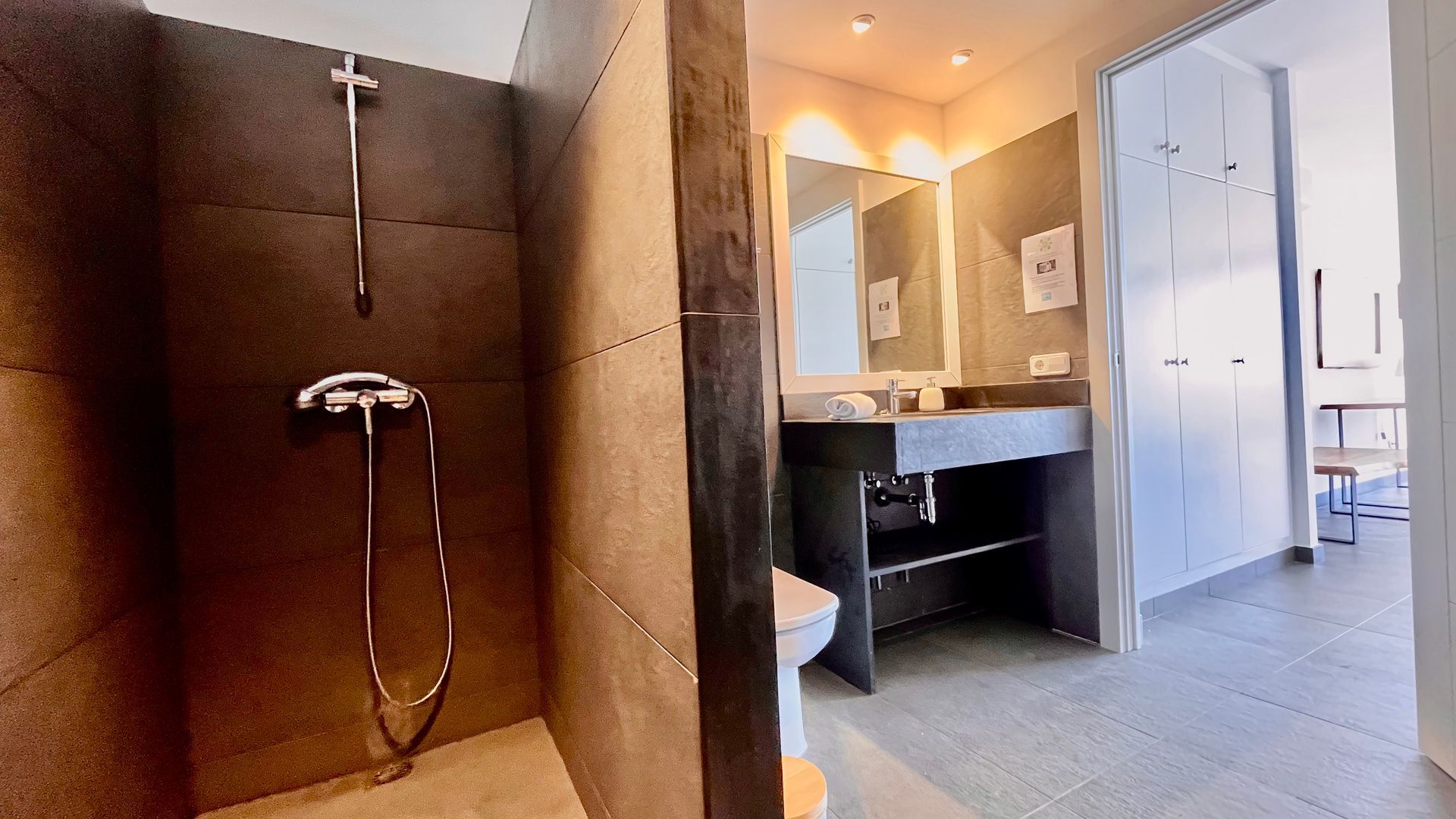 Baño moderno con ducha a ras de suelo, lavabo e inodoro. Paredes oscuras, tonos neutros. Luz brillante.