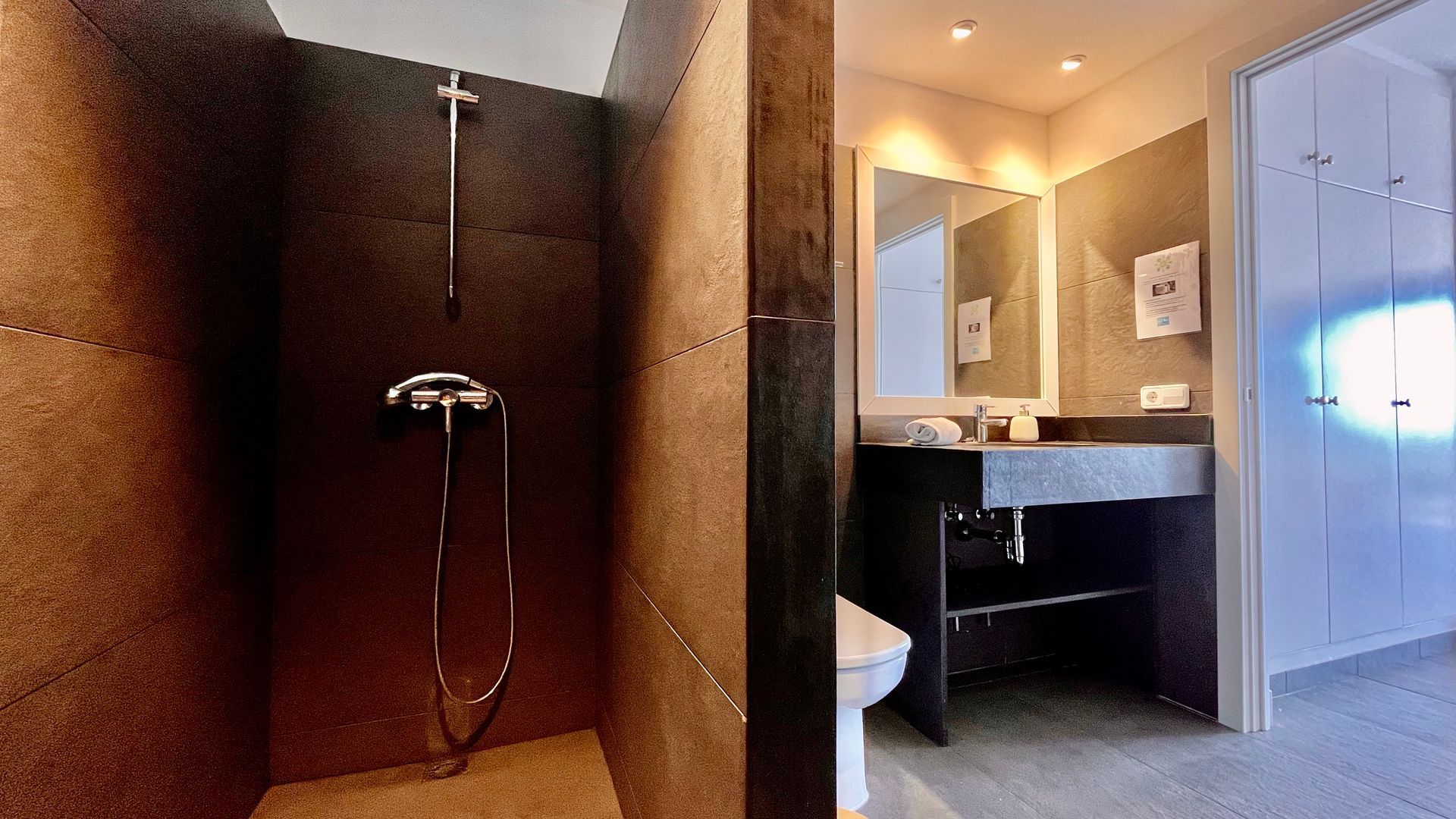 Baño moderno con ducha y tocador con azulejos oscuros, paredes blancas y armario.