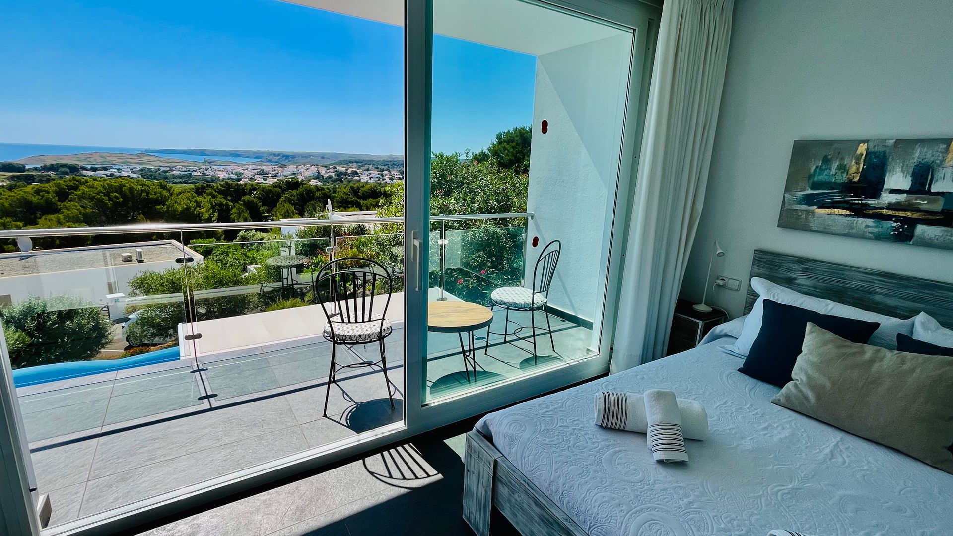 Dormitorio con balcón con vistas a la ciudad y a los árboles. Decoración en blanco y azul, cama, silla y mesa pequeña.