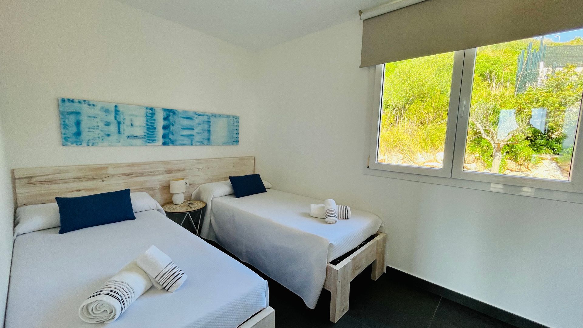 Dormitorio doble con camas blancas, detalles en azul y una ventana con vista a la vegetación.