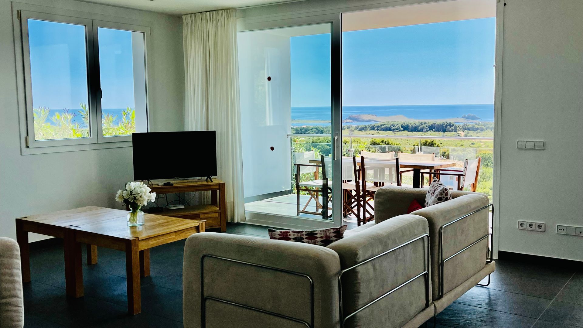 Sala de estar con vista al mar: sofá de color claro, mesa de madera, TV, puerta corrediza abierta a un patio.