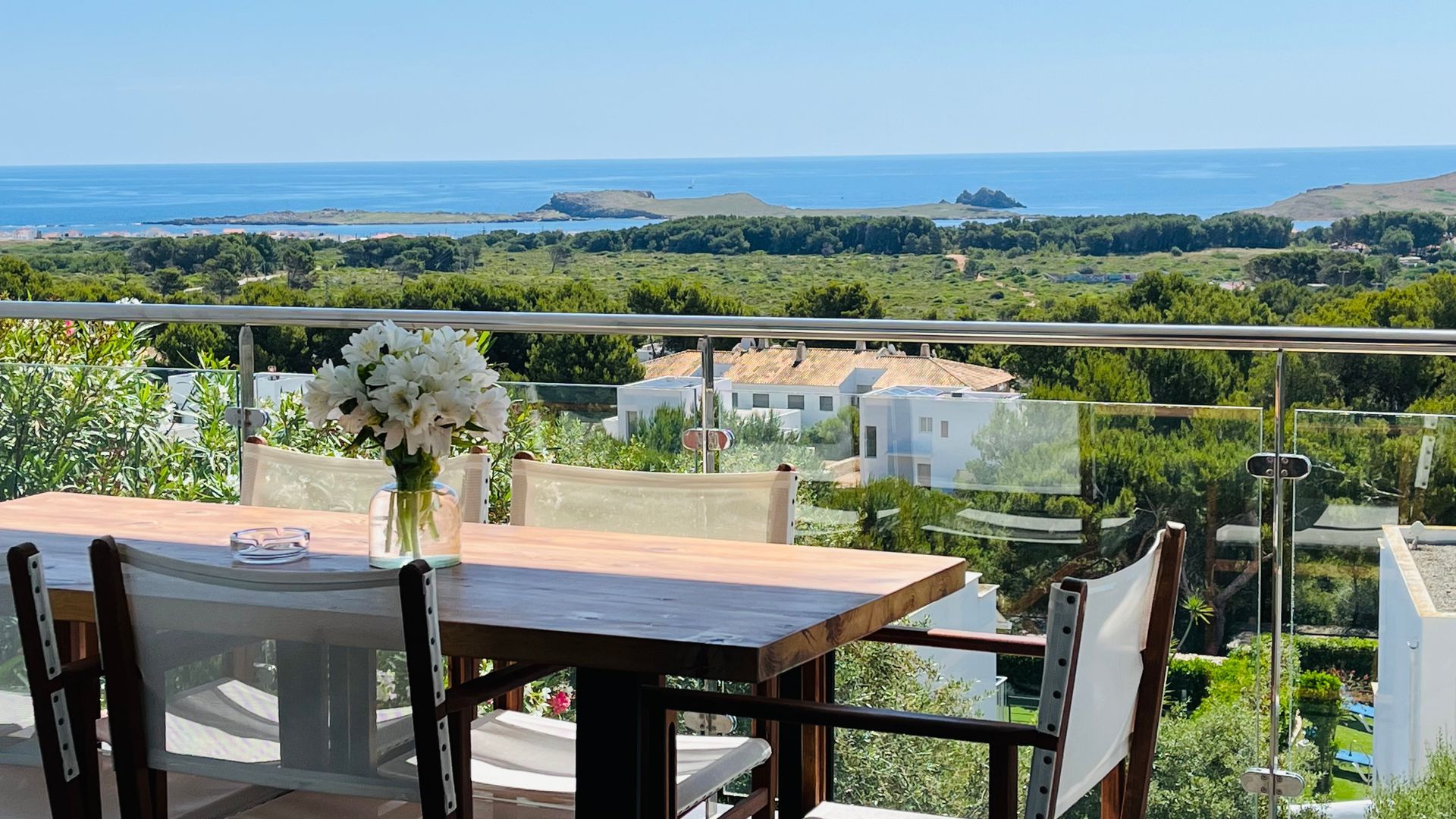 Balcón con mesa, sillas, flores, vista al mar, árboles y edificios en un día soleado.