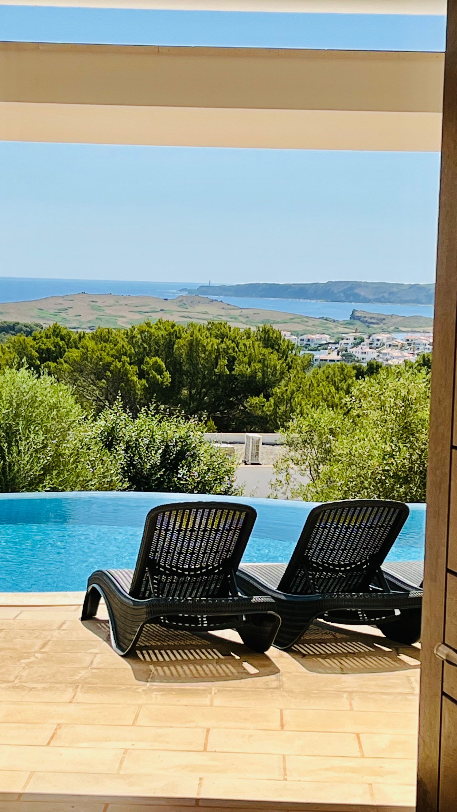 Dos tumbonas negras junto a una piscina con vistas a un paisaje y al océano bajo un cielo azul.