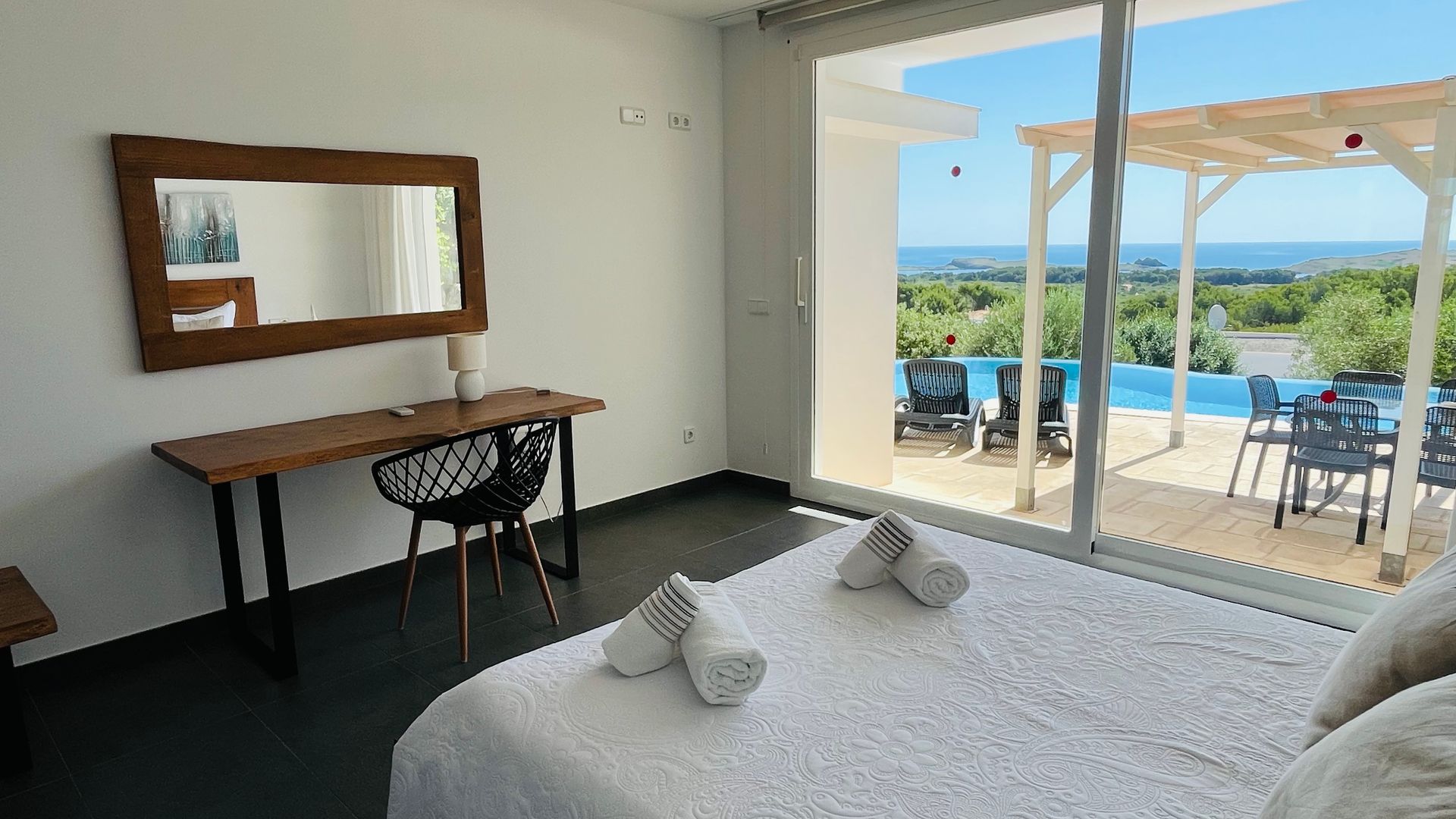 Dormitorio con cama, escritorio y puerta corrediza a terraza con vista a la piscina y al mar.