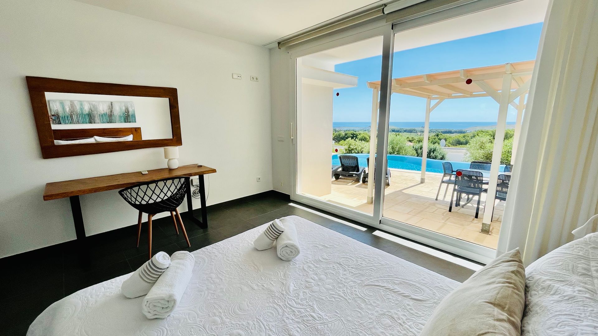 Dormitorio con vista a la piscina y al mar. Ropa de cama blanca, escritorio de madera y grandes puertas de cristal.