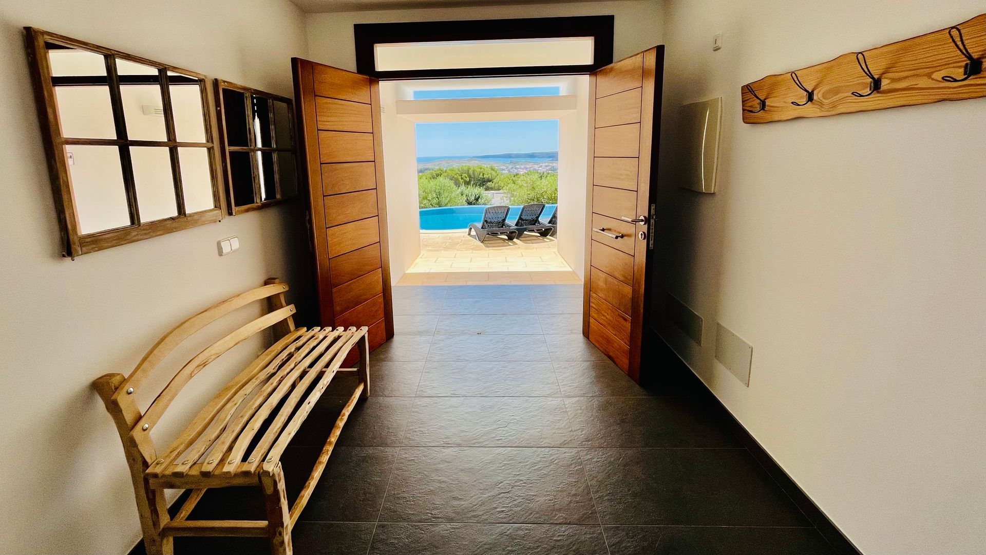 Pasillo con puertas dobles abiertas que dan a la piscina exterior de agua azul. Incluye banco de madera y perchero.