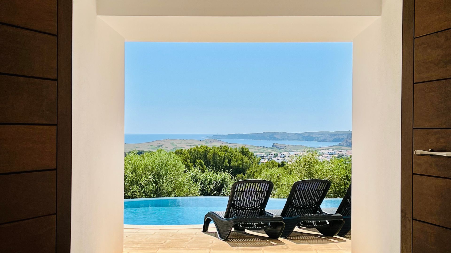 Vista a través de una puerta de una piscina y tumbonas con vistas a un paisaje con cielo azul.