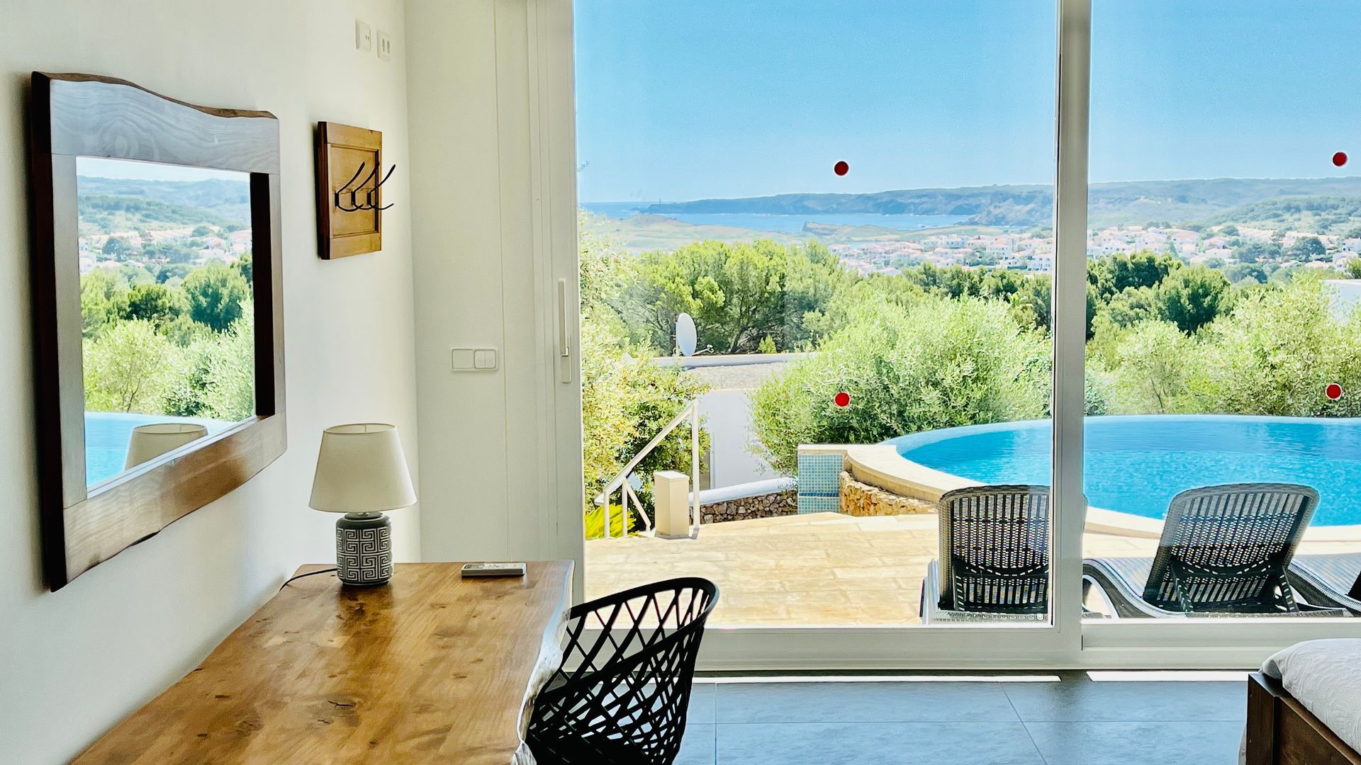 Dormitorio con escritorio, espejo y puerta abierta a una piscina con vista a un paisaje.