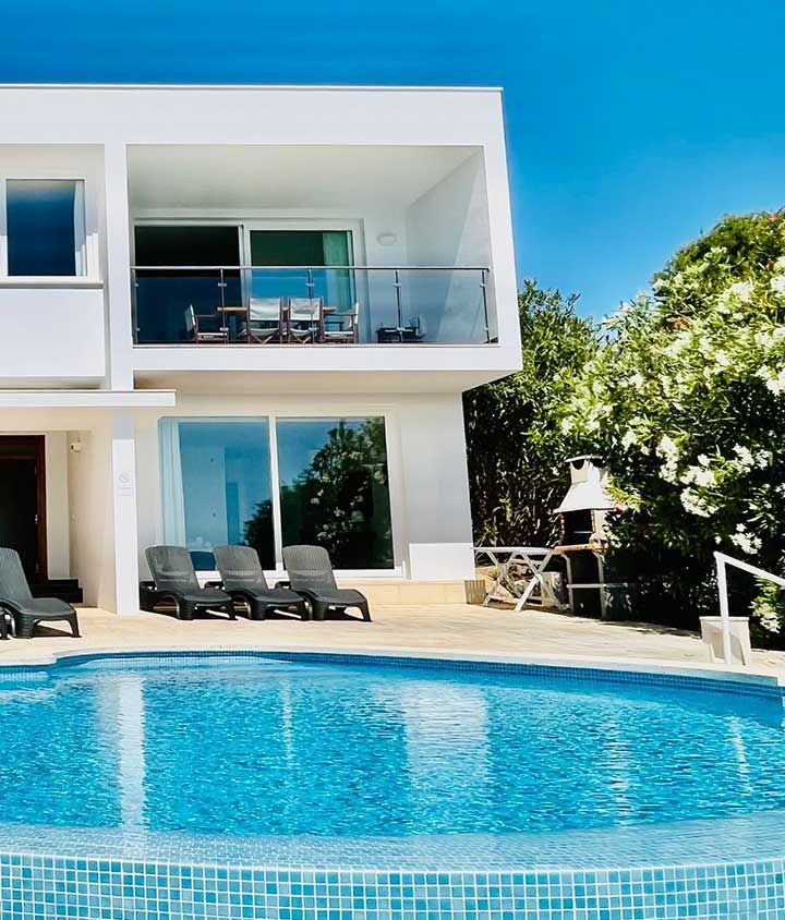 Moderna villa blanca con piscina, tumbonas y vista al mar.