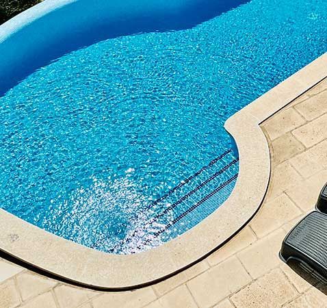 Escena junto a la piscina con agua azul, piedra beige y tres tumbonas grises.