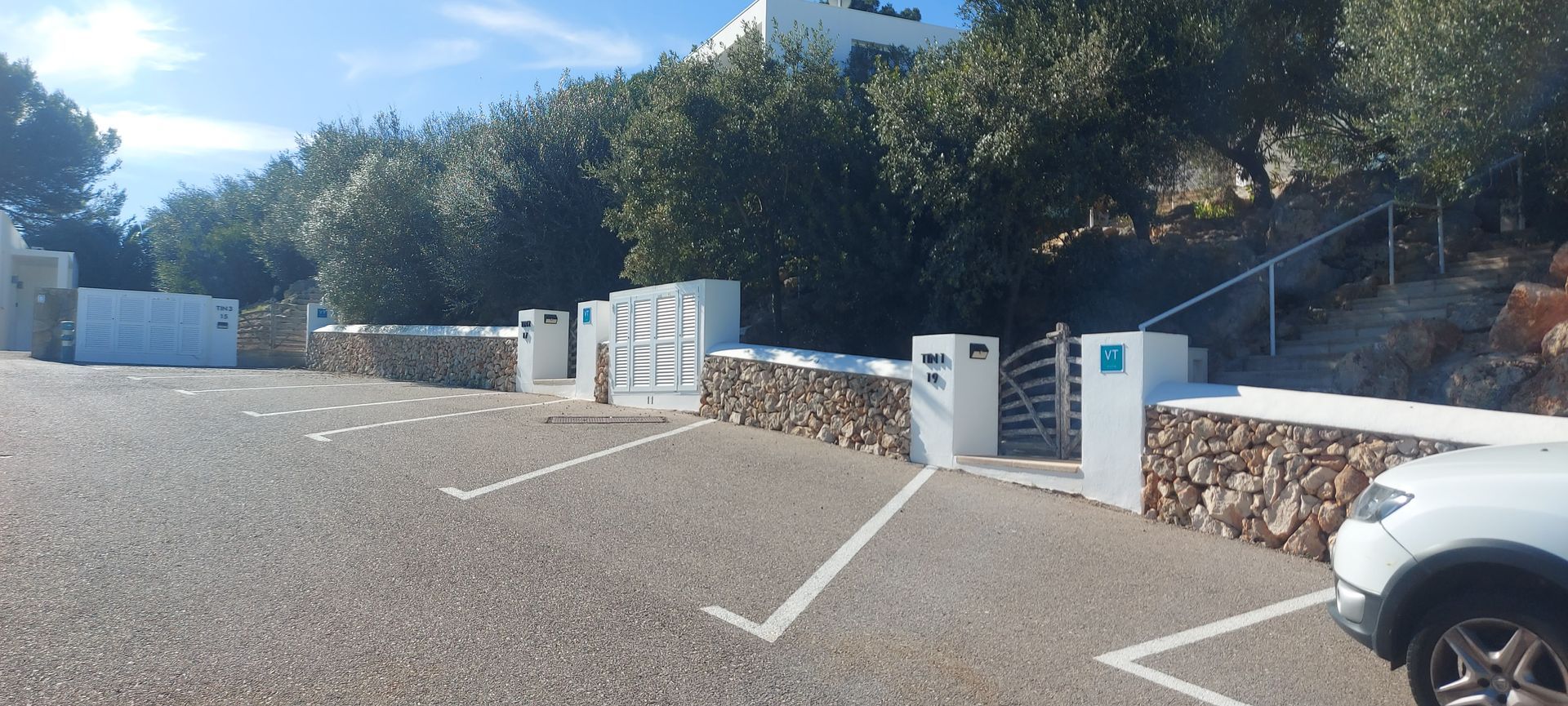 Zona de aparcamiento con coche blanco aparcado delante de un muro de piedra con portón de entrada y arbolado.