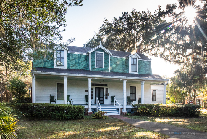 A white house with a green roof and a porch surrounded by trees.