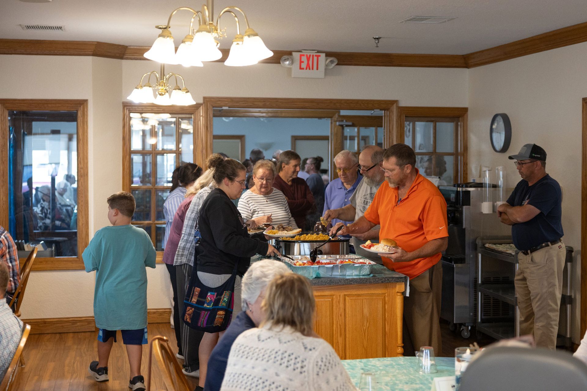 People serving food from a buffet in a dining room. Some stand in line, others seated.