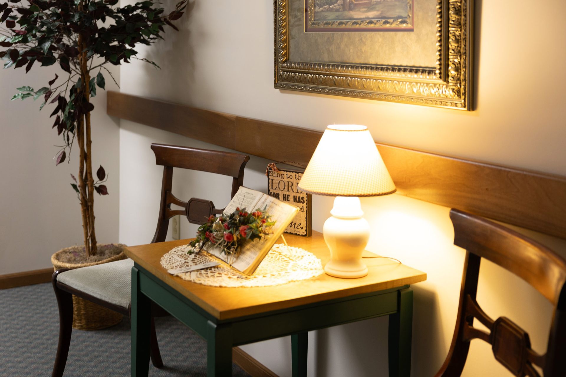 Cozy corner: Table with lamp, open book with flowers, next to a tree and chairs, under a framed picture.