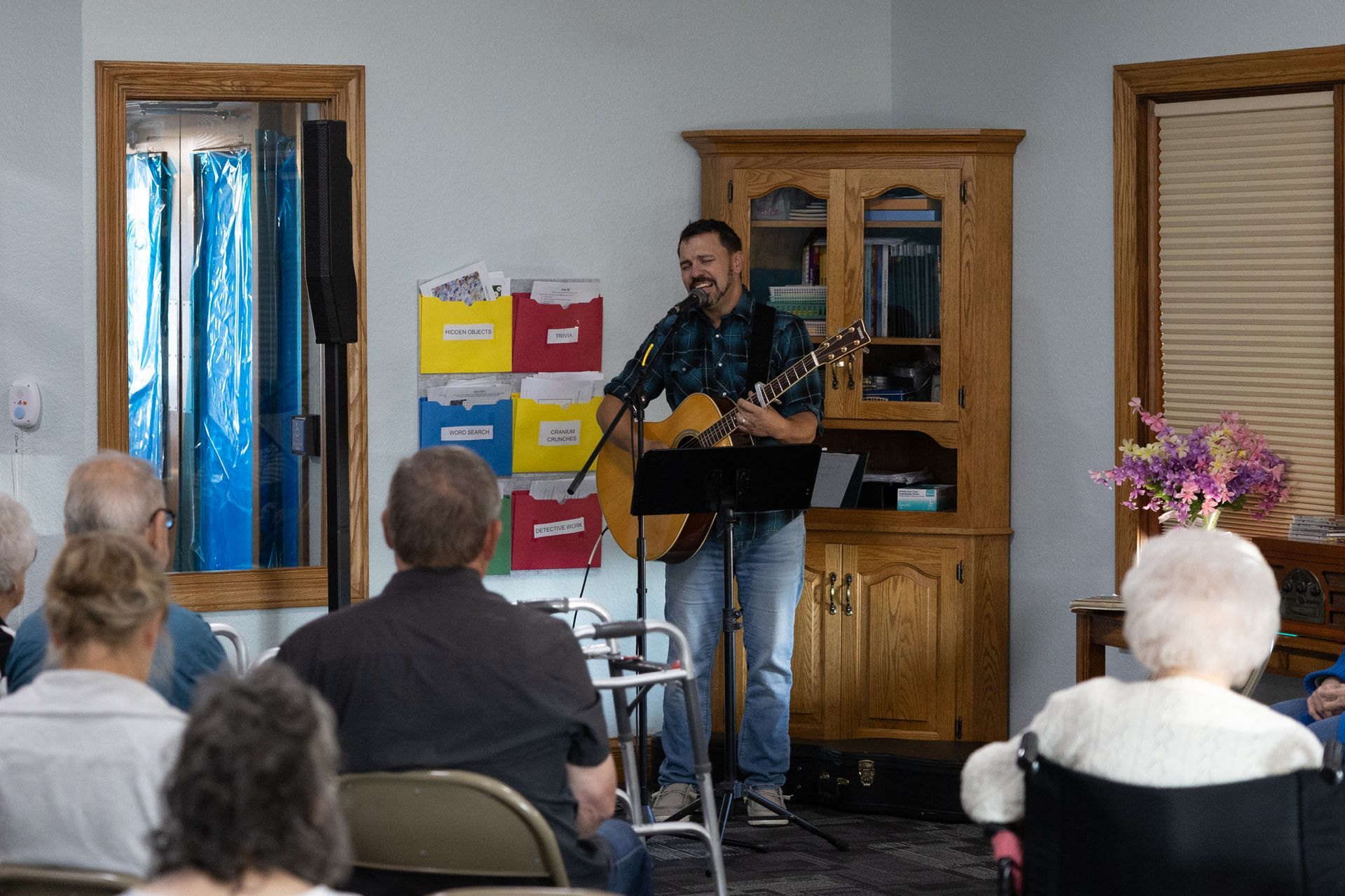 A man plays guitar for a seated audience in a room, likely a senior center.