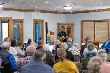 A man plays guitar for an audience in a bright room with windows and a wooden cabinet.