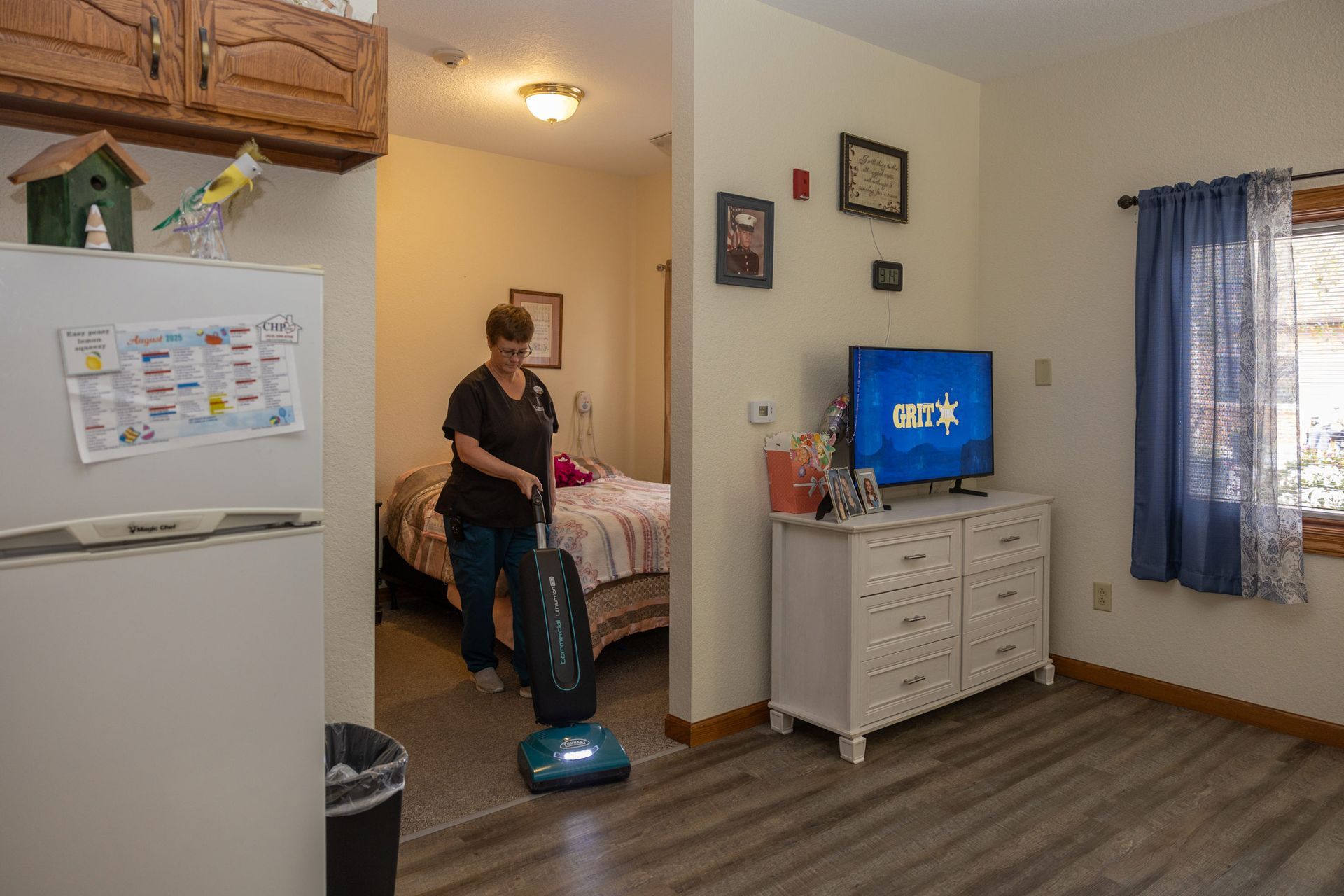 Woman vacuums a room with a bed, dresser, and a window.