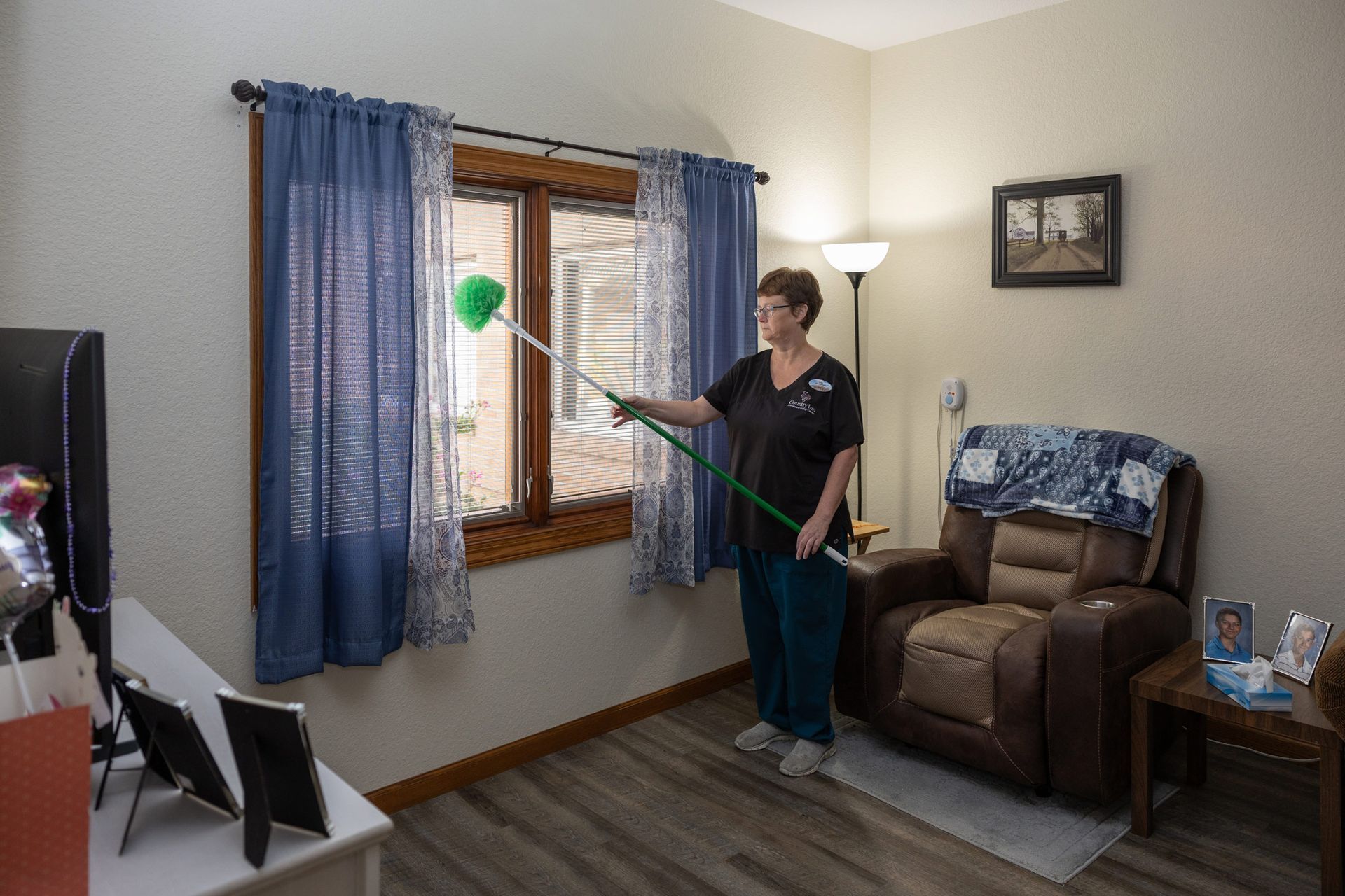 Woman dusting a window with blue curtains. Inside a room with a recliner and a floor lamp.