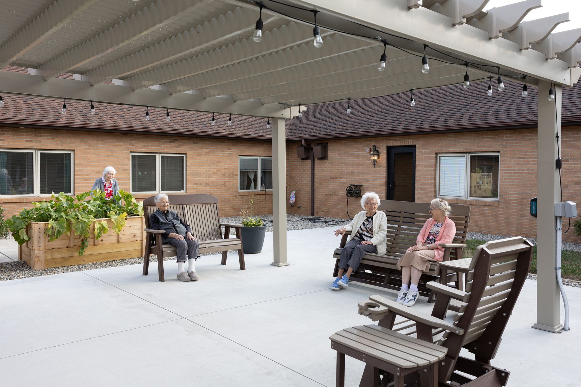Seniors relax on benches under a pergola near a brick building.