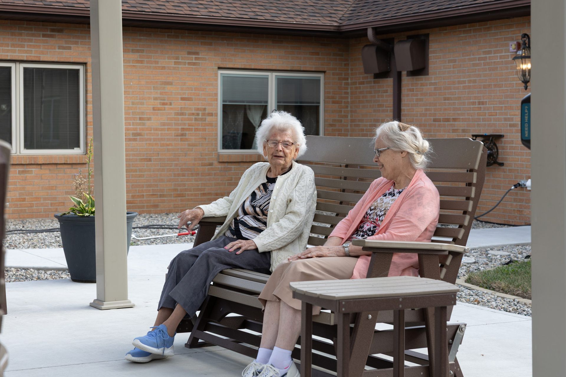 Two elderly women chat on a porch swing, smiling. Brick building background, neutral colors.
