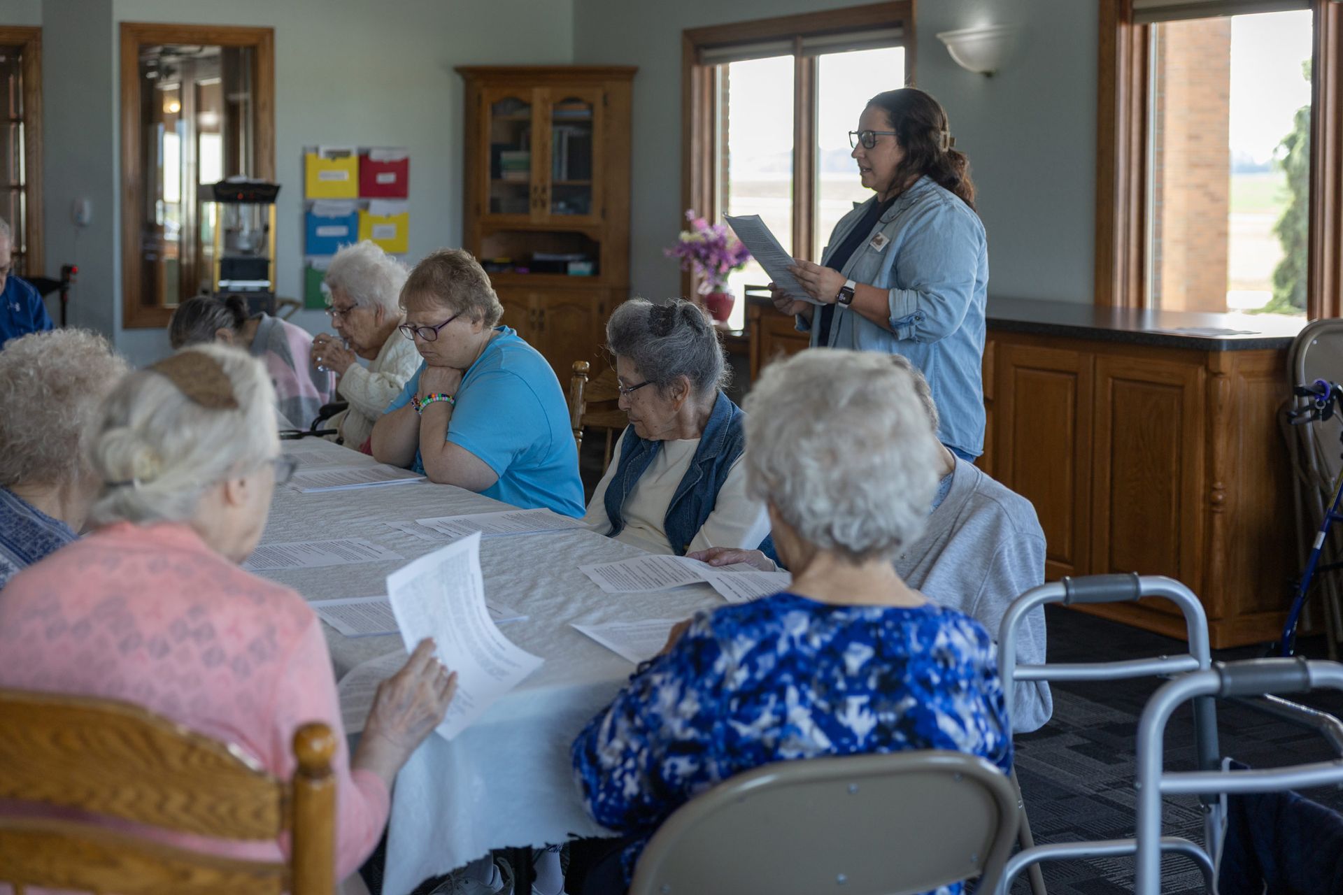 A group of elderly women in a room listening to a woman reading from a tablet.