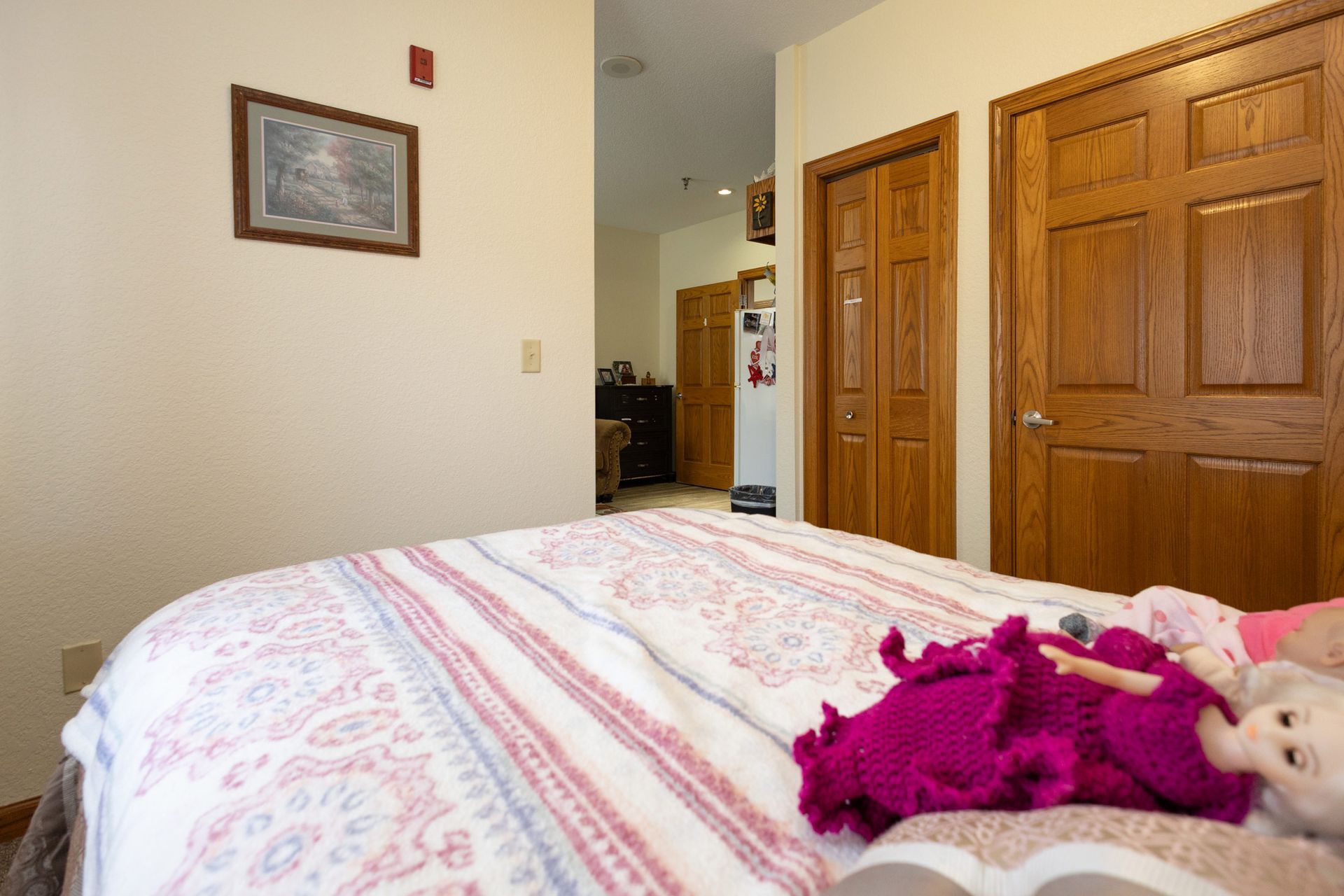 Bedroom with bed, two dolls, two wooden doors, and an open doorway to a hallway.