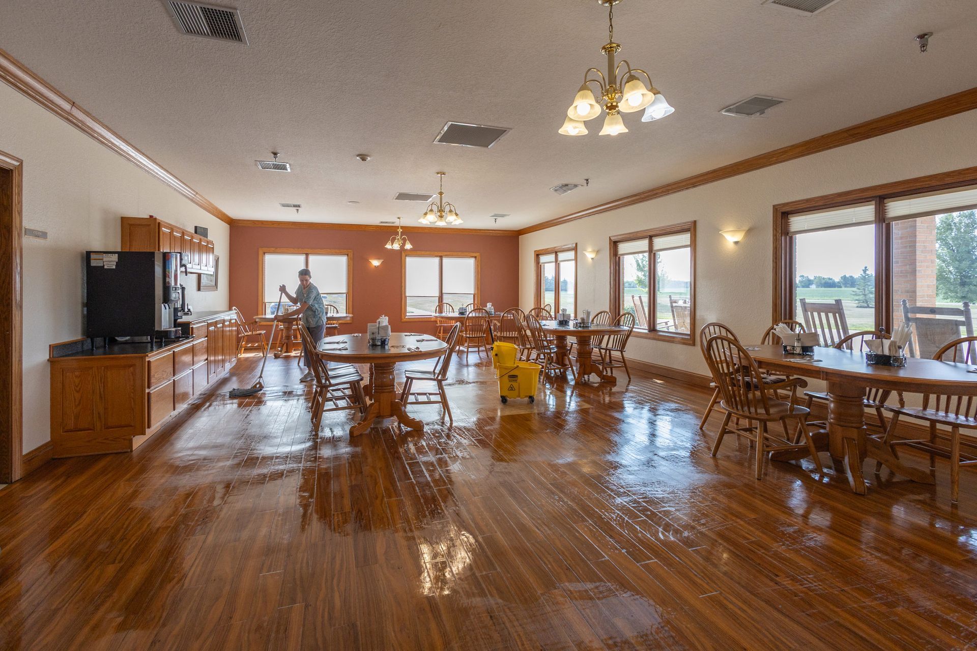 Woman mopping a shiny wooden floor in a large room with round tables, chairs, and windows.