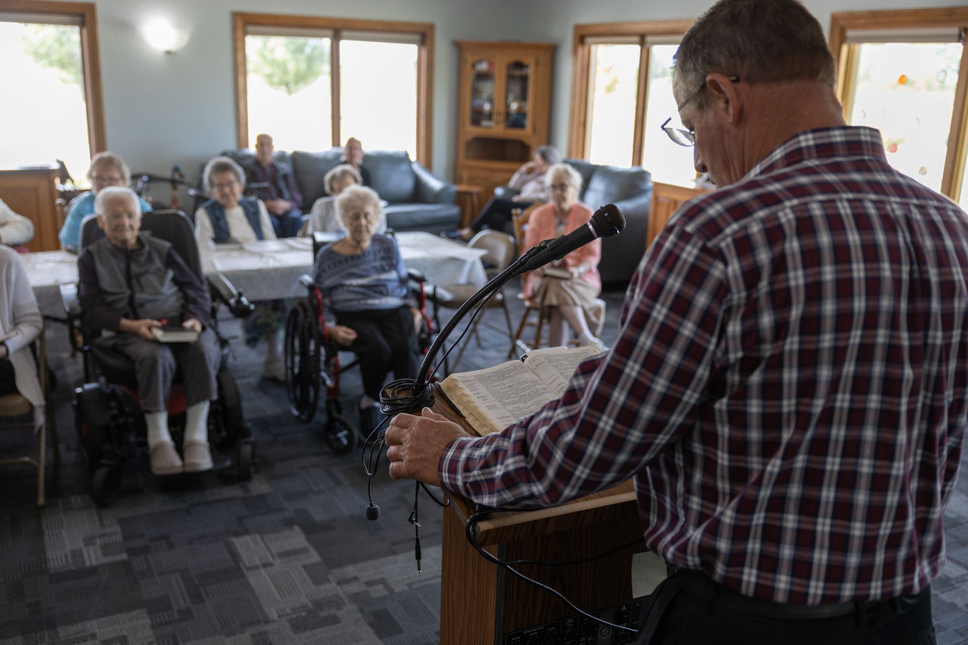 Man reading at a podium to seated elderly people in a room with tables and chairs.