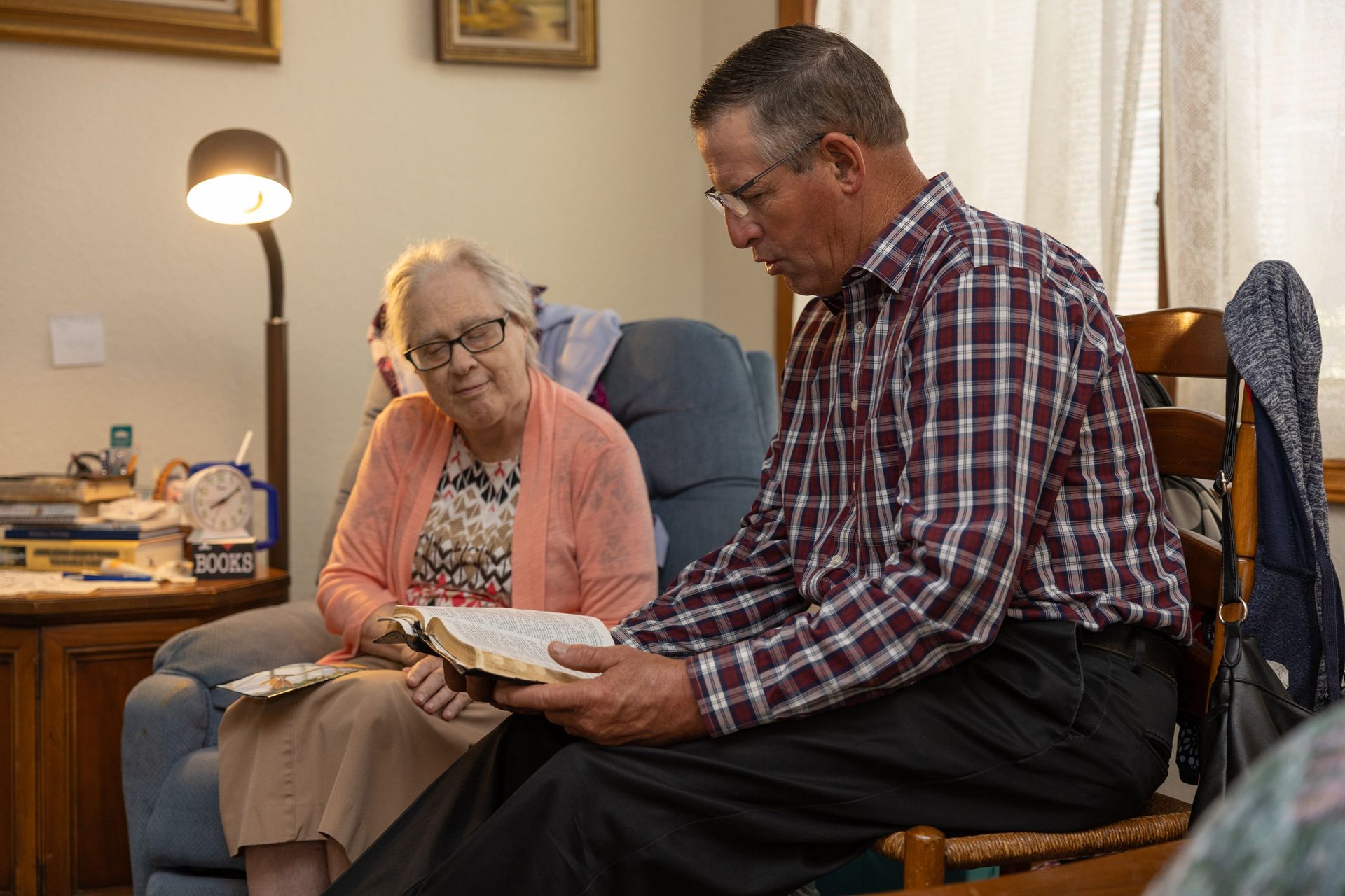 An older man reads a book to an older woman in a living room, both seated.