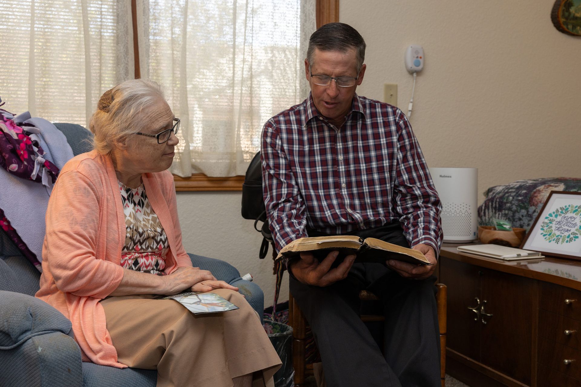 An elderly woman and a man reading from a Bible together, sitting in a living room.