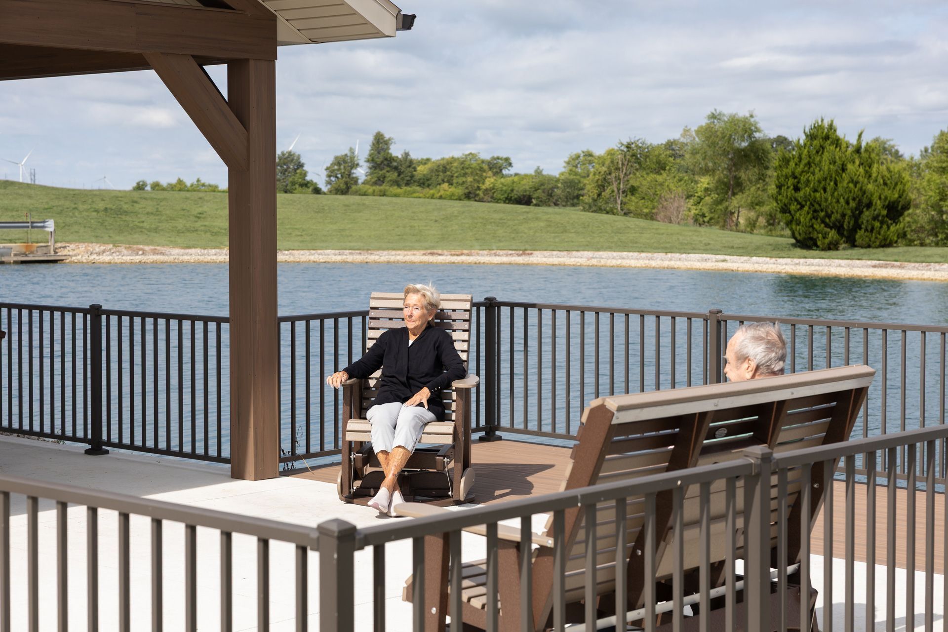 Two seniors relax on a patio overlooking a lake, under a wooden gazebo.