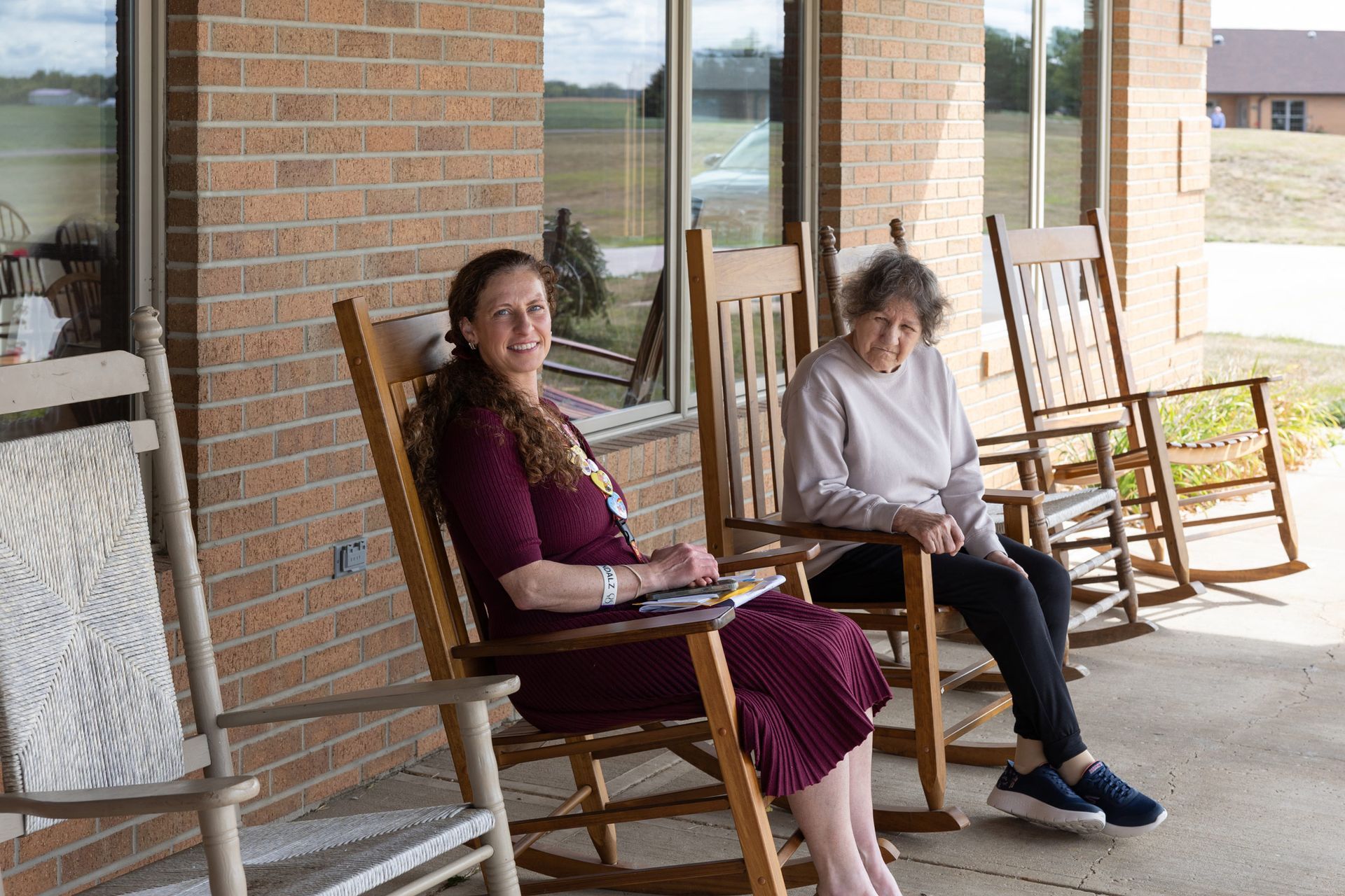 Two women in rocking chairs on a porch; one smiles, the other looks forward. Sunny day, brick building.