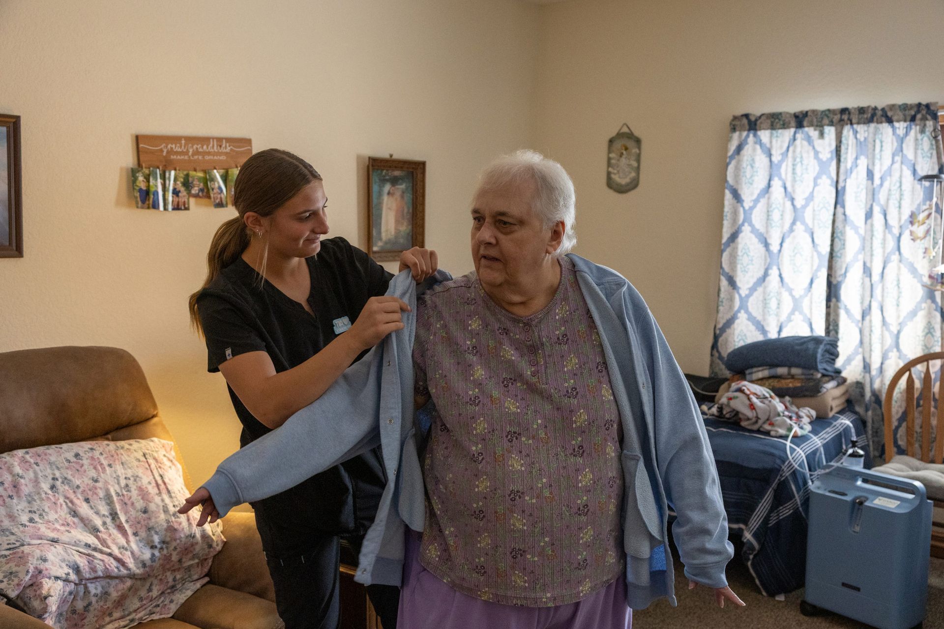 A caregiver helps an elderly woman put on a cardigan in a home setting.