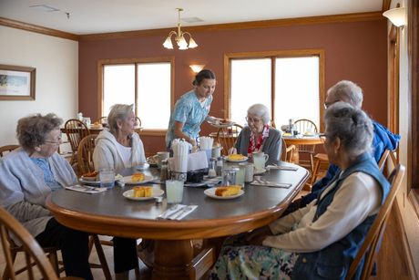 A server serves breakfast to a group of senior citizens seated at a round table in a dining room.