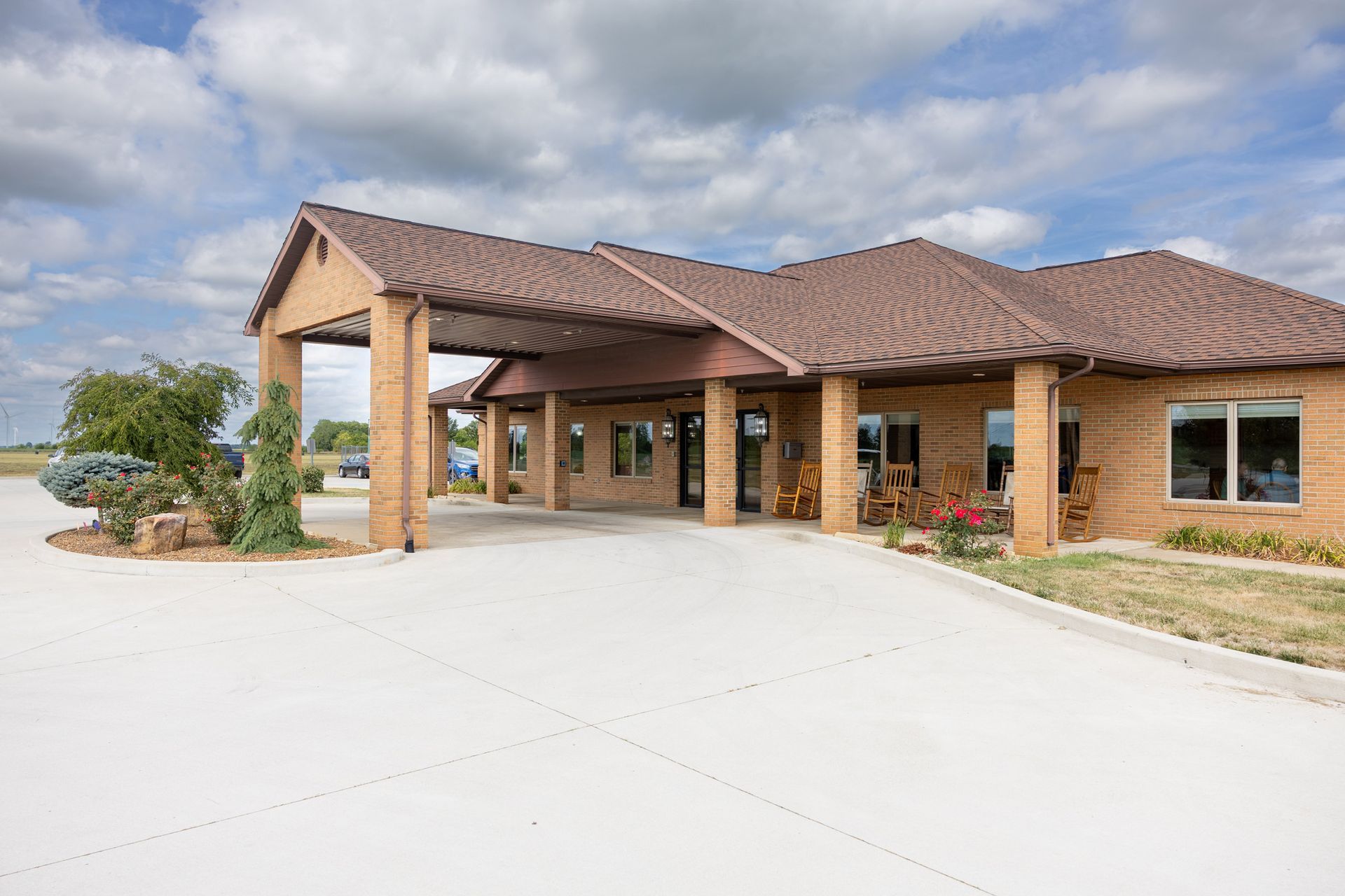 Tan brick building with brown roof and covered entrance. Concrete driveway, blue sky.