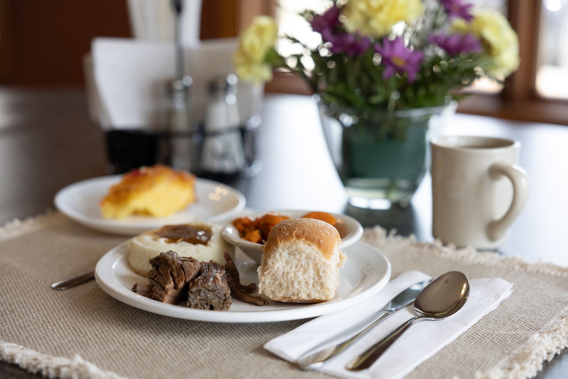 Plate of food with meat, bread, mashed potatoes, and sweet potatoes, with coffee, flowers.