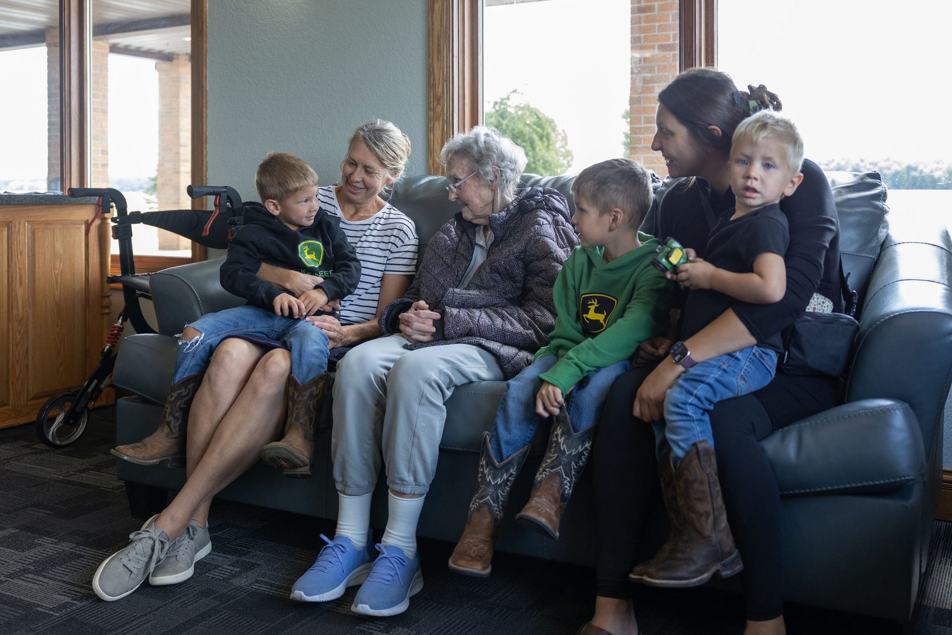 Family group sits on a couch. Two women, two children, and an older woman smiling. Daylight setting.