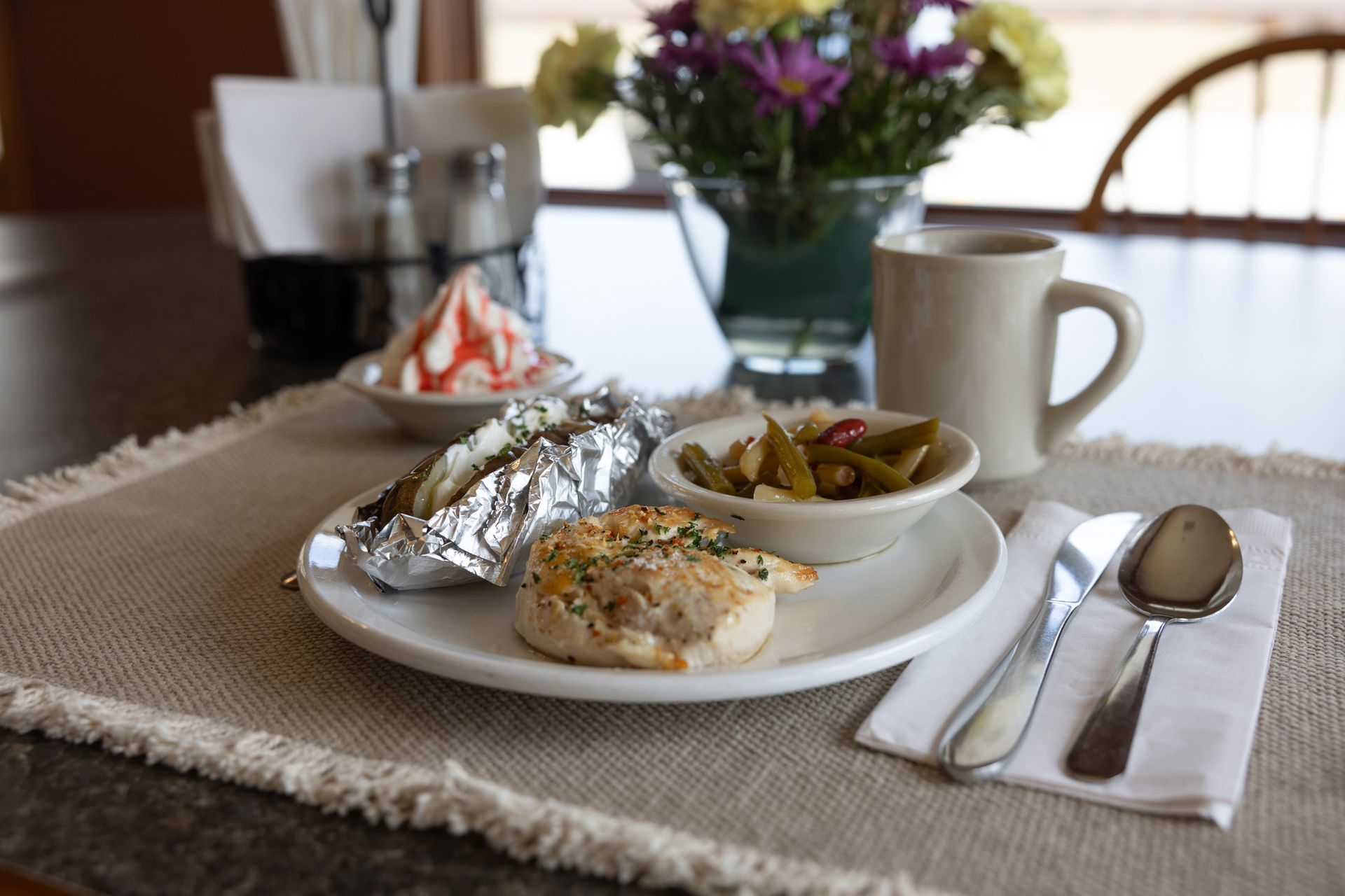 Meal on a table: baked potato, roll, green beans, dessert, coffee, flowers, silverware.