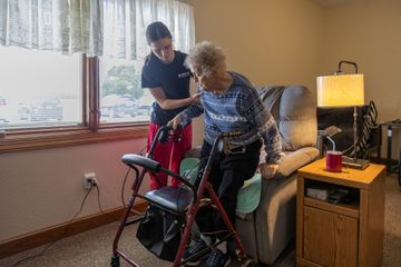 Caregiver assists elderly woman with walker to stand from recliner. Indoor, warm lighting.