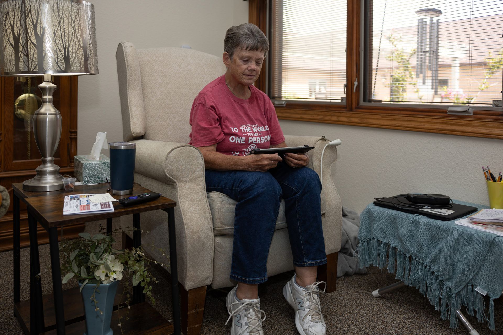 Woman in a red shirt sits in a chair, looking at a tablet. Next to a window, a lamp, and side table.
