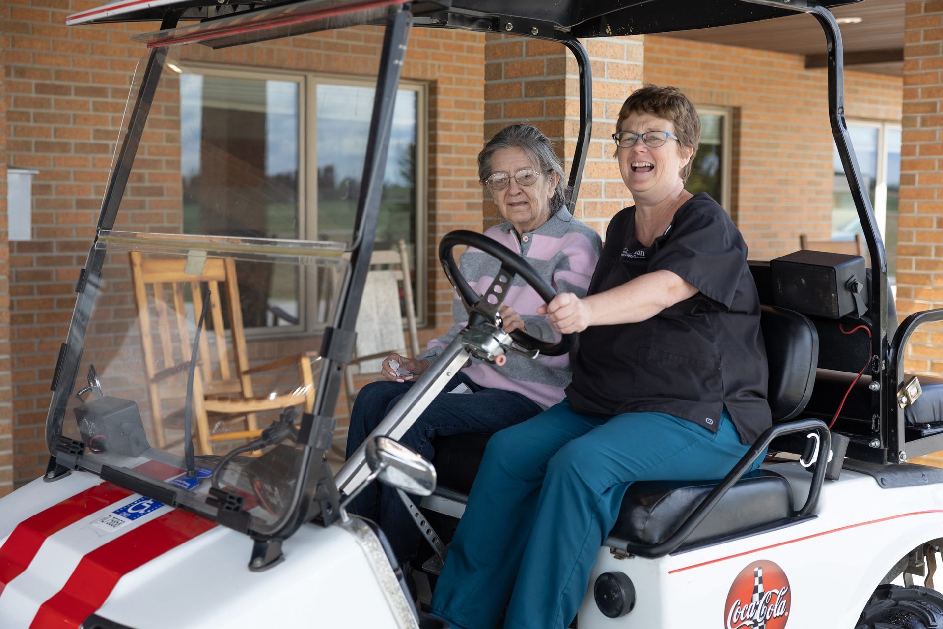 Two women smile in a white golf cart decorated with red, white, and blue stripes. Brick building in background.