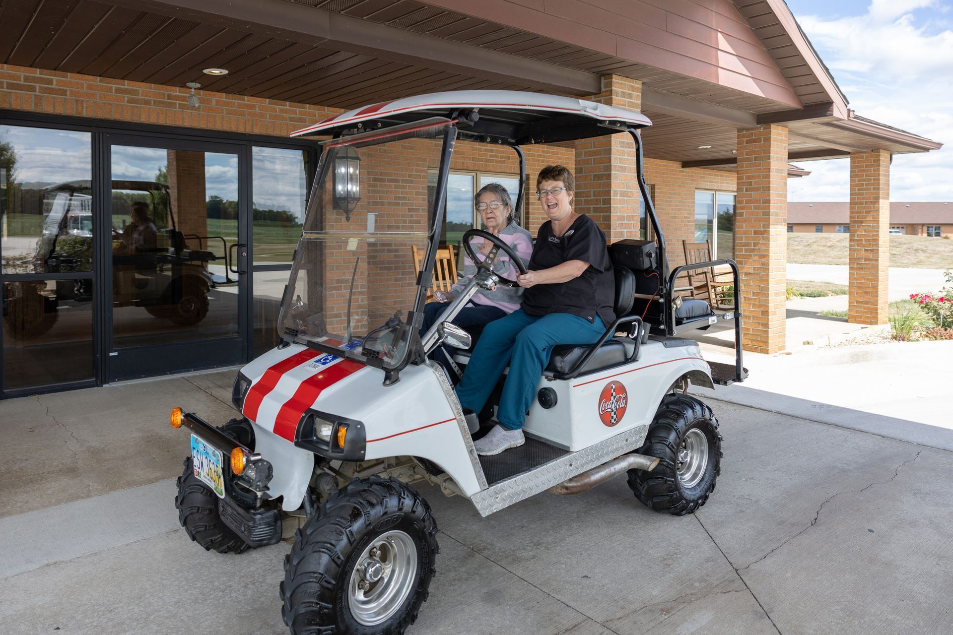 Two women in a golf cart decorated with red stripes and a cross. The cart is outside a building.