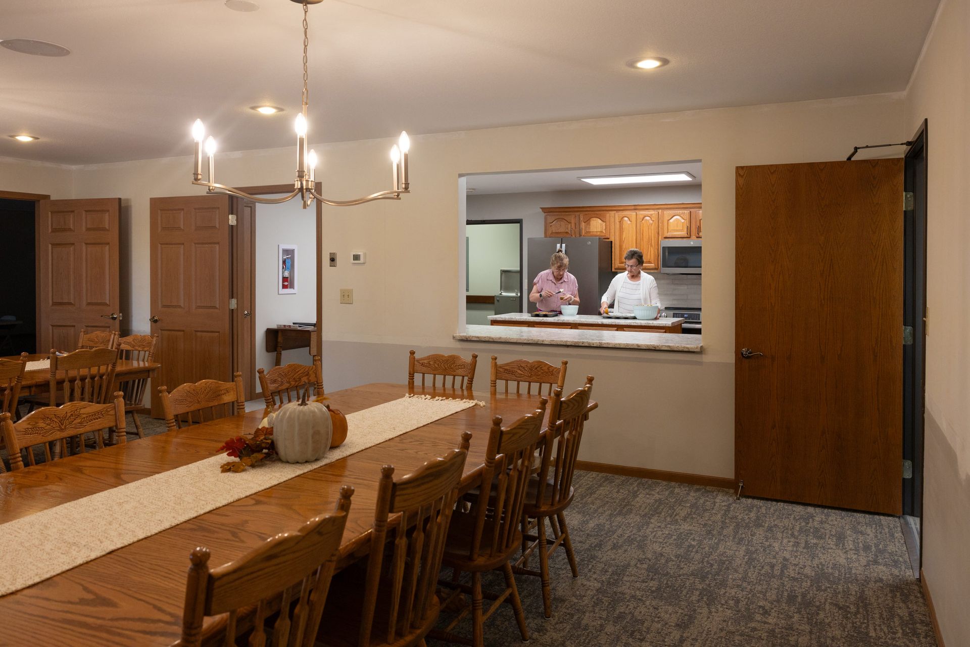 Dining room with long wooden table, people in kitchen pass-through.