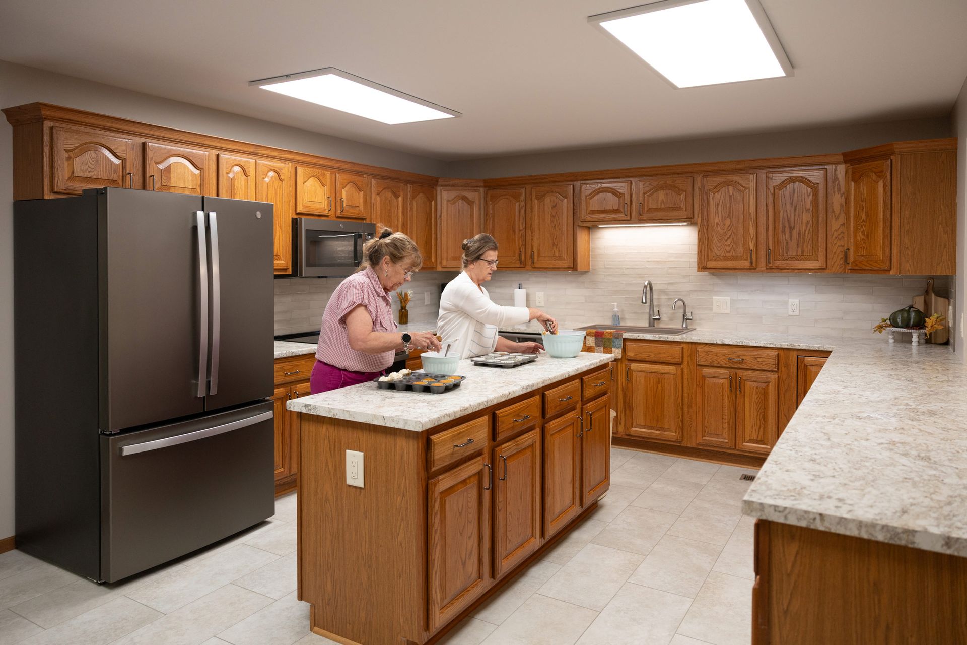 Two women bake in a kitchen with oak cabinets, stainless steel appliances, and an island.