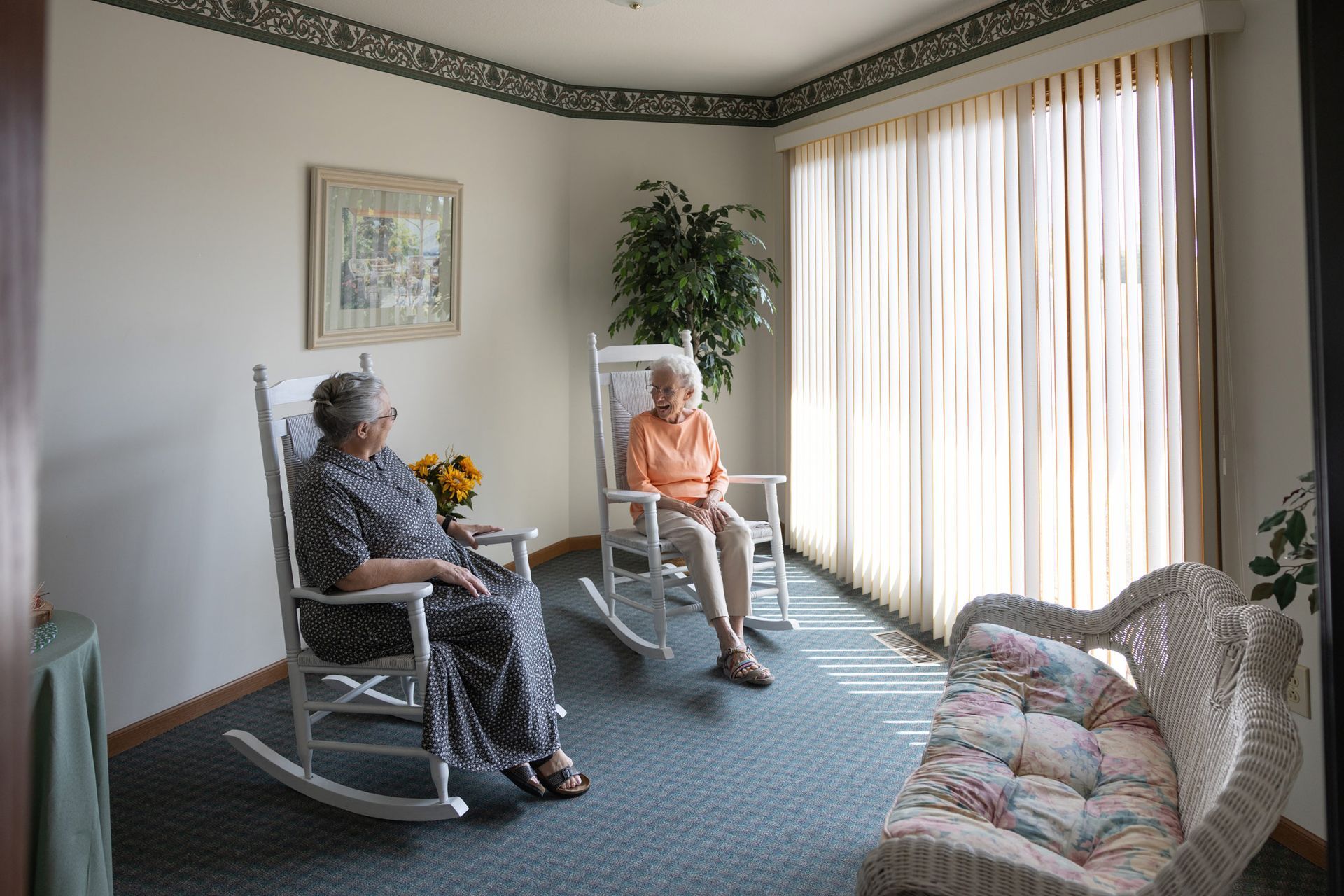 Two older women sit in rocking chairs in a bright room with curtains. One has a floral dress; the other, an orange shirt.
