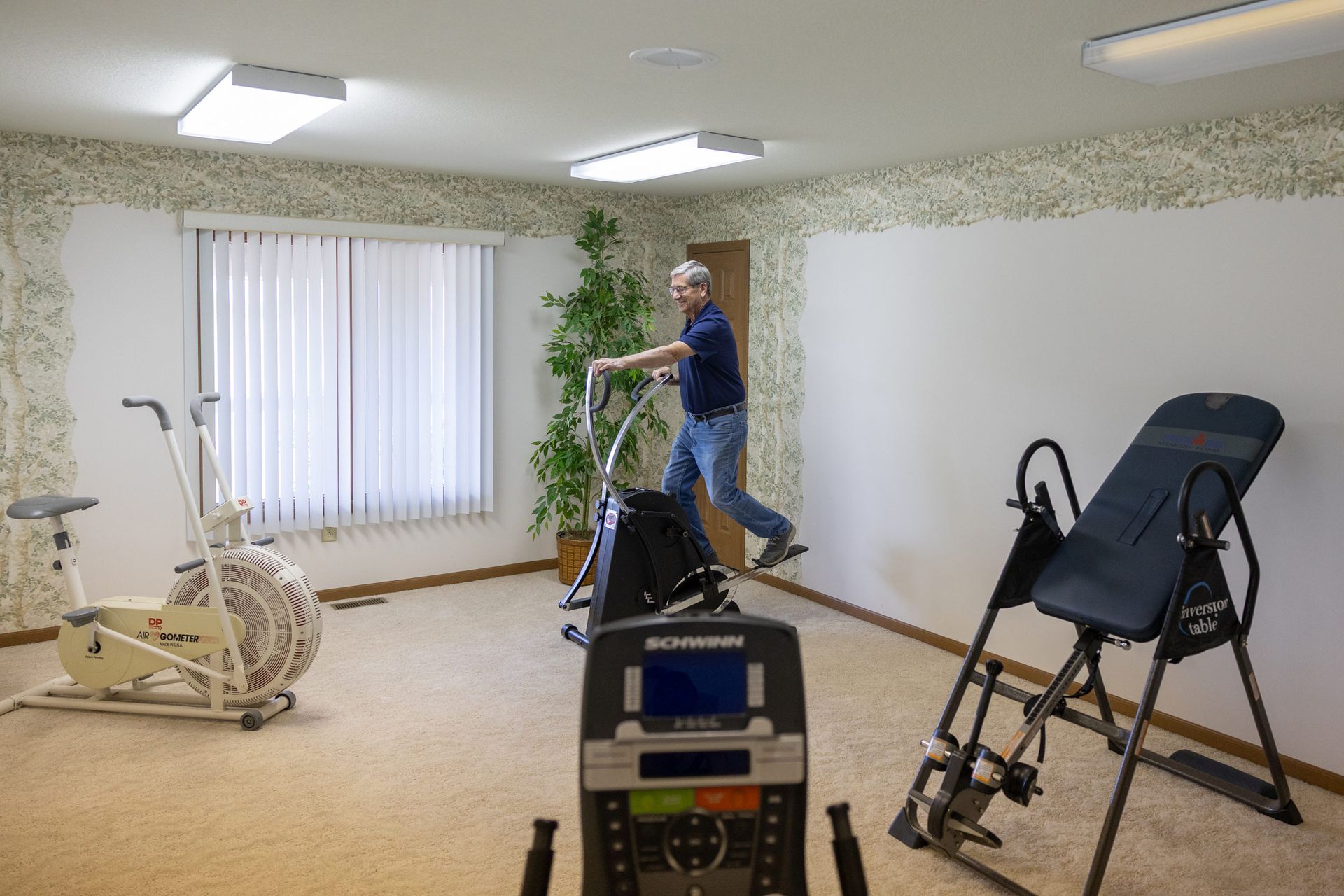 Man exercising in a home gym with an elliptical, inversion table, and a stair climber.