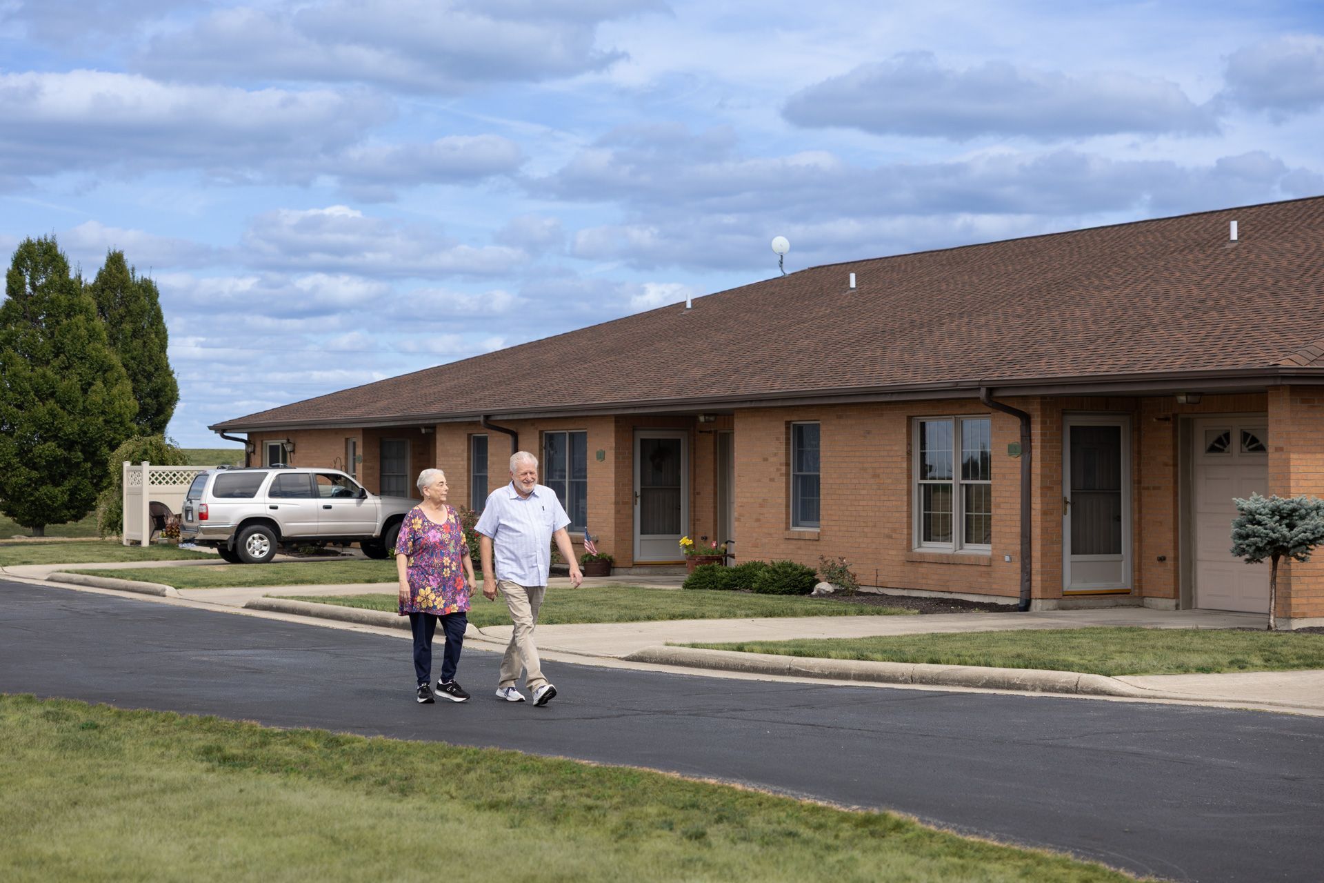 Elderly couple walking on a paved path in front of brick apartments on a sunny day.