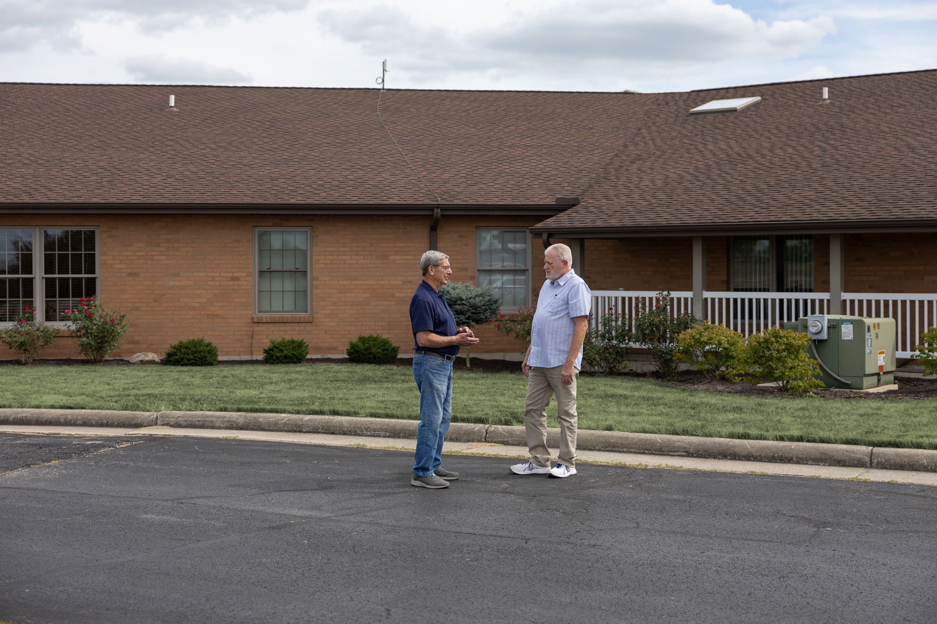 Two men standing on pavement in front of a brick building, conversing outdoors.