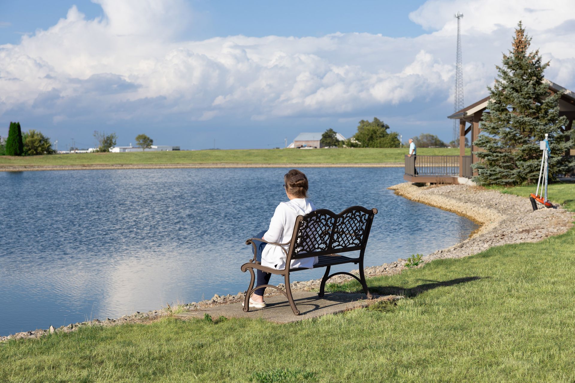 Person sitting on bench, gazing at calm lake under a cloudy sky. Lakeside building and trees visible.