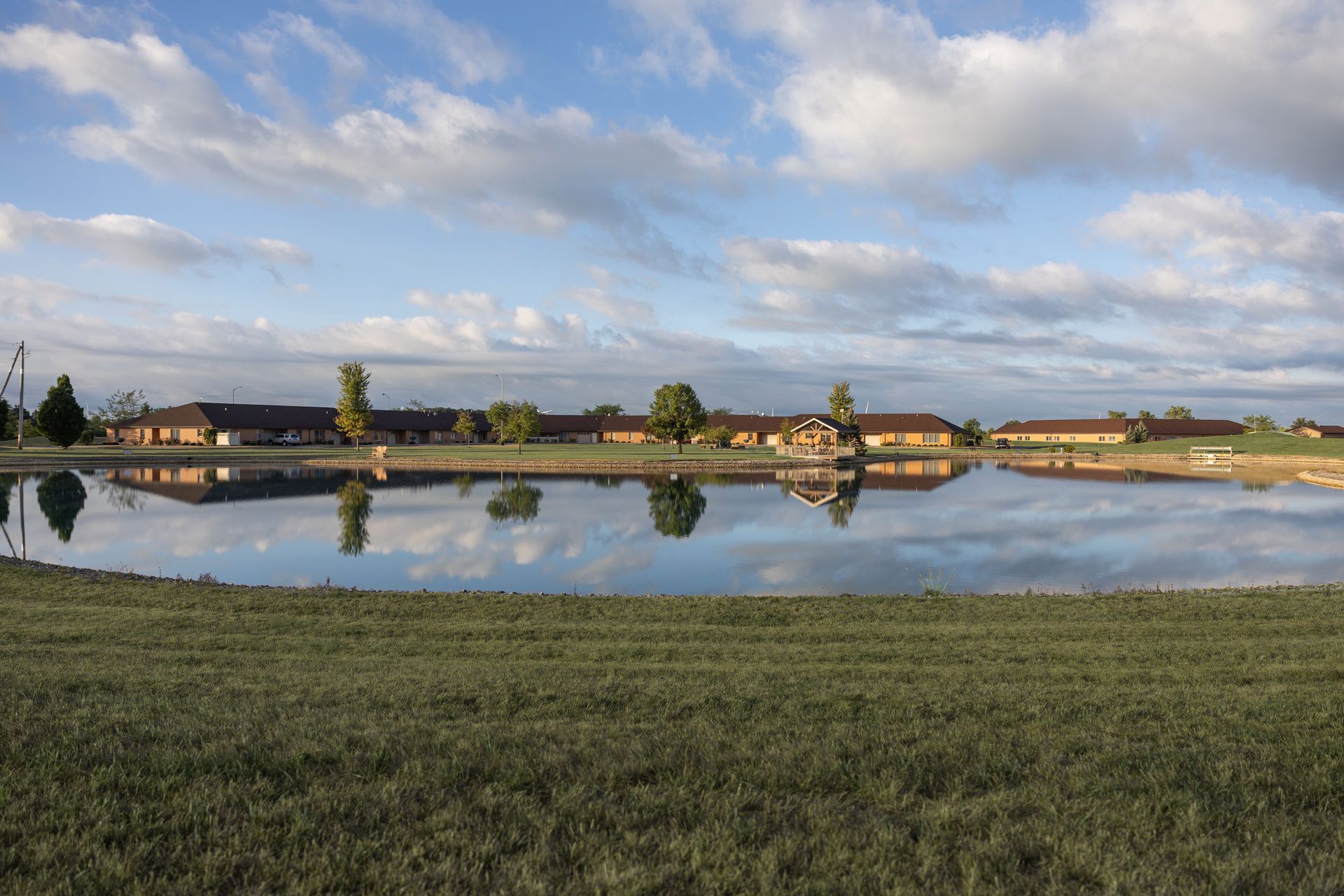 Buildings reflected in a calm lake under a cloudy sky, with dry, golden field in foreground.